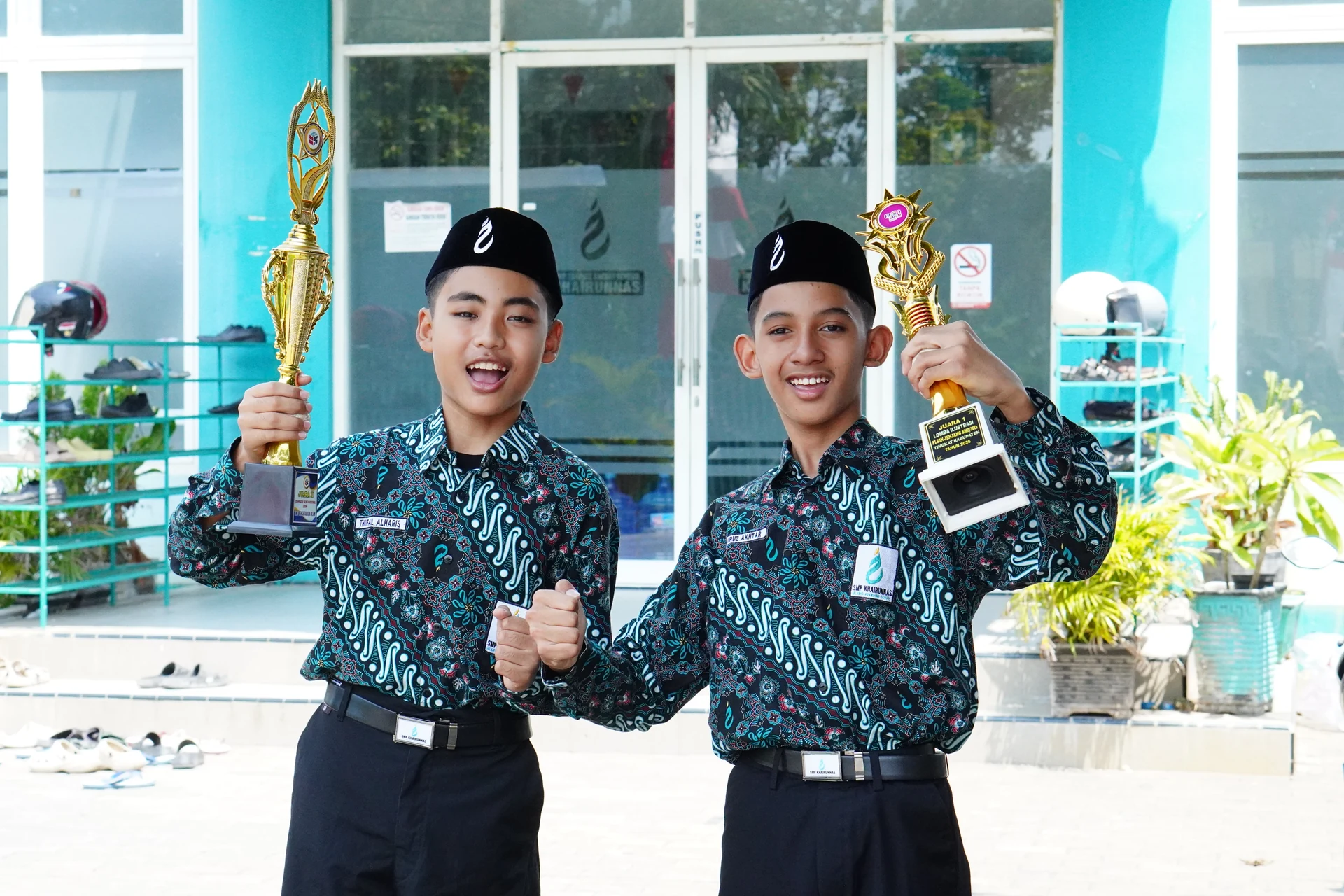 Dua santri putra berpose dengan piala di depan gedung sekolah.
