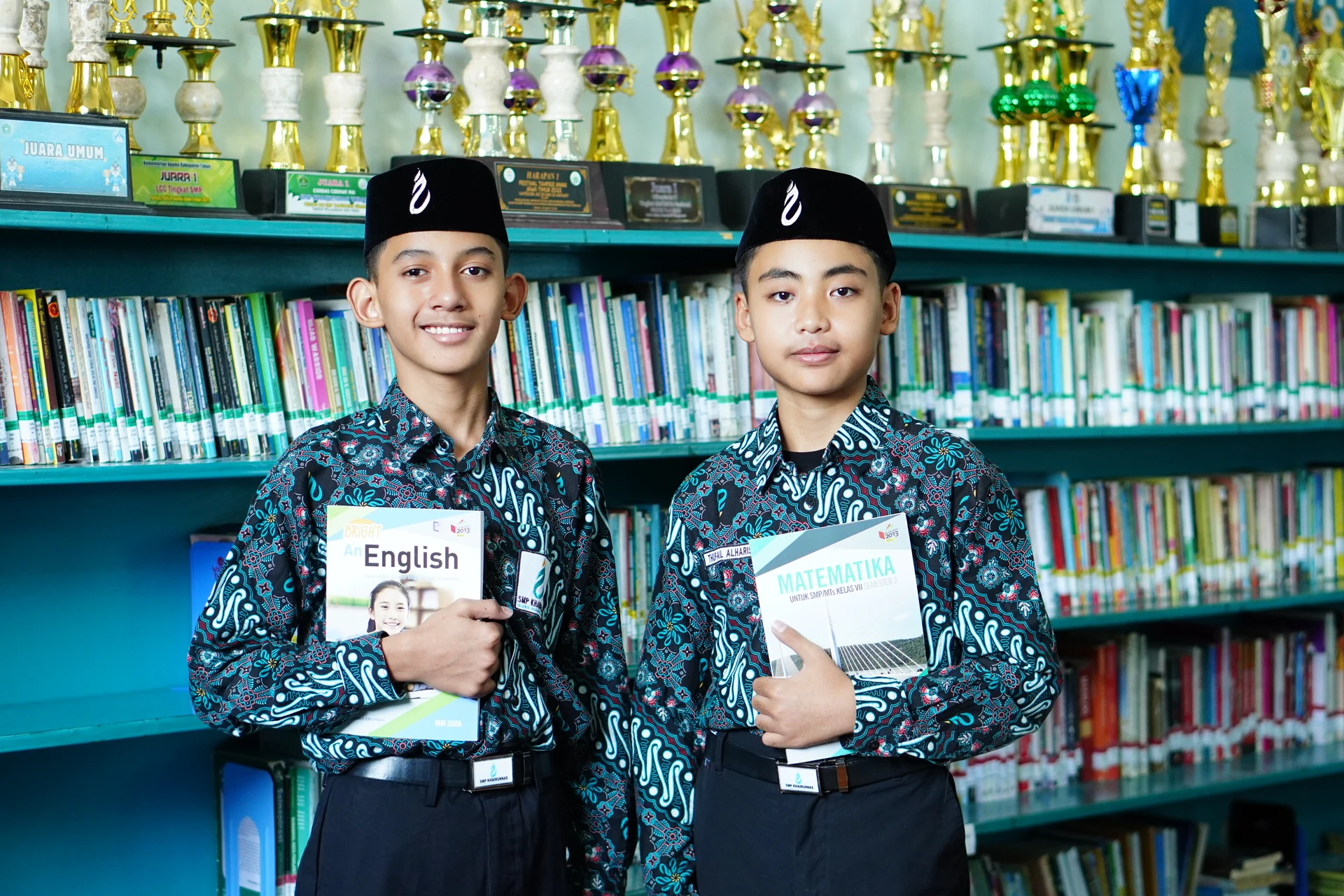 Dua santri putra membawa buku di depan rak perpustakaan.